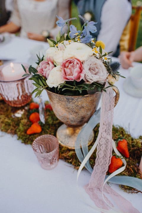 Hochzeitswahn - Ein rustikaler Tafelaufsatz bestehend aus einer Vintage-Trophäenvase mit rosigen Rosen und Wildblumen, akzentuiert durch zarte Spitze, auf einem Tischläufer aus Moos bei einer Veranstaltung im Freien.