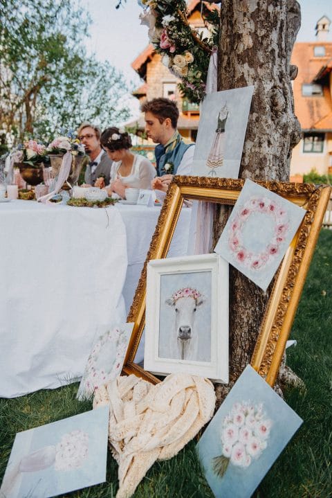Hochzeitswahn - Eleganter Hochzeitsempfang im Freien mit Gästen, die eine Mahlzeit genießen, umgeben von kunstvoll arrangierten, skurrilen Gemälden und verzierten goldenen Rahmen, die an einem Baum lehnen.