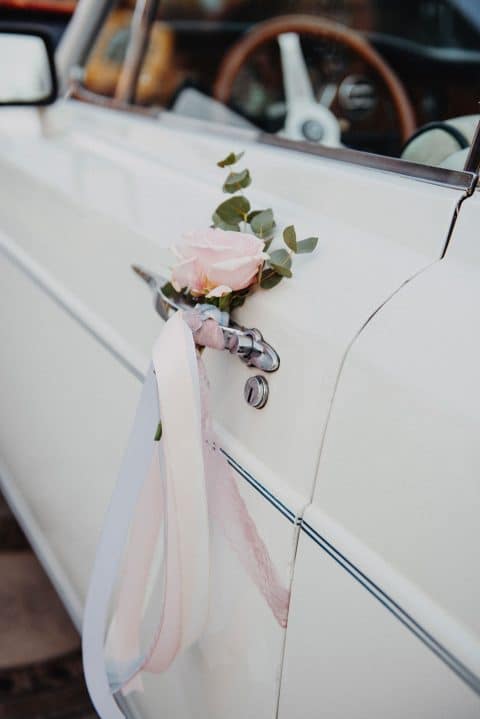 Hochzeitswahn - Ein weißes Vintage-Hochzeitsauto, geschmückt mit einer zarten rosa Rose und Grünpflanzen und befestigt mit weichen Bändern, vermittelt ein Gefühl romantischer Eleganz und zeitloser Tradition.