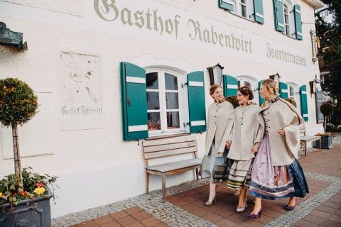 Hochzeitswahn - Drei Personen in traditioneller bayerischer Kleidung gehen lachend vor dem „Gasthof Rabenwirt“, einem malerischen Gebäude mit grünen Fensterläden, die die lokale deutsche Kultur und Gastfreundschaft symbolisieren.
