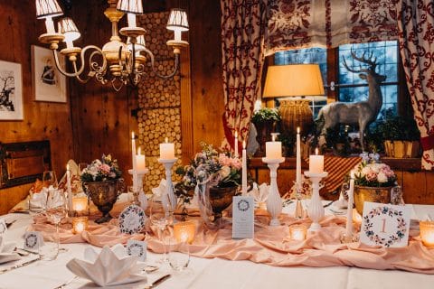 Hochzeitswahn - Ein elegant gedeckter Esstisch mit Blumenschmuck, Kerzen und einem rustikalen Kronleuchter schafft eine warme Atmosphäre in einem holzgetäfelten Raum mit gemütlichen Vorhängen und Kunstwerken.