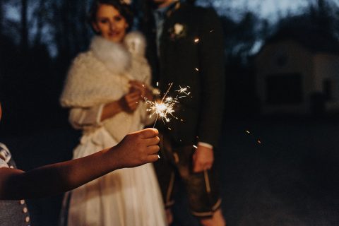 Hochzeitswahn - Ein Kind streckt einer lächelnden Braut und einem Bräutigam in festlicher Kleidung eine Wunderkerze entgegen, die gemeinsam einen freudigen Abendmoment feiern.