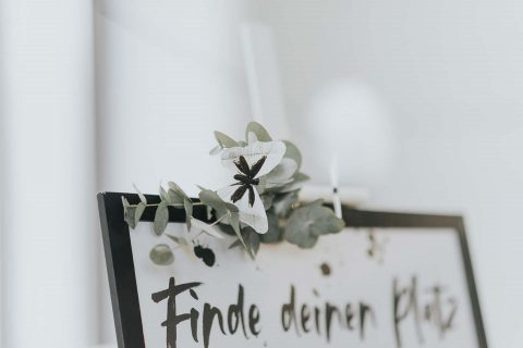 Hochzeitswahn - Ein zartes Blumenarrangement ziert ein Schild mit dem deutschen Satz „Finde deinen Platz“, der in einer legeren, handgebürsteten Schrift geschrieben ist und eine einladende Stimmung hervorruft, die oft bei Versammlungen oder Veranstaltungen verwendet wird.