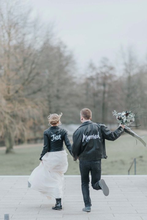 Hochzeitswahn - Ein fröhliches Brautpaar, gekleidet in Lederjacken mit der Aufschrift „Just married“, spazieren Hand in Hand, die Braut hält einen Blumenstrauß in der Hand, und feiern ihre Verbindung mit einer skurrilen Note vor der Kulisse eines ruhigen Parks.