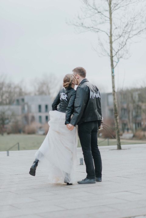 Hochzeitswahn - Ein Paar im Hochzeitskleid, mit personalisierten Jacken, auf denen stolz „frisch verheiratet“ steht, teilt einen zärtlichen Moment vor einer städtischen Kulisse, während die Zukunft ihnen so offen steht wie der Himmel über ihnen.