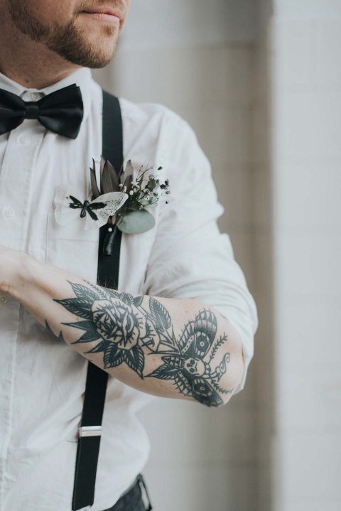 Hochzeitswahn - Eine Person in Abendgarderobe mit Fliege und Hosenträgern, die ein detailliertes Blumentattoo auf dem Unterarm präsentiert, ergänzt durch eine schicke Boutonniere am Revers.