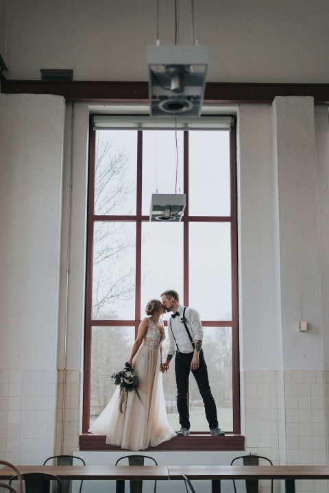 Hochzeitswahn - Ein Paar im Hochzeitskleid teilt einen zärtlichen Moment auf einer Bank neben einem hohen Fenster in einem Interieur im Industrie-Chic-Stil im sanften Tageslicht.