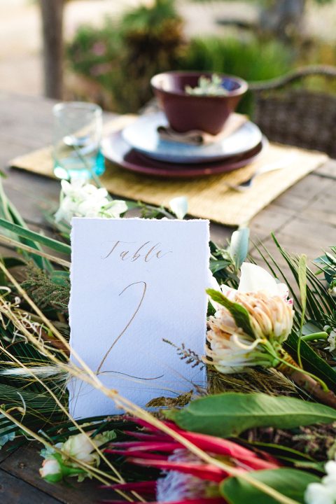 Hochzeitswahn - Ein eleganter Hochzeitstisch im Freien verfügt über eine handgeschriebene Tischnummer inmitten eines üppig grünen Tischaufsatzes, mit feinem Geschirr und Gläsern, die für eine anspruchsvolle Feier bereitstehen.