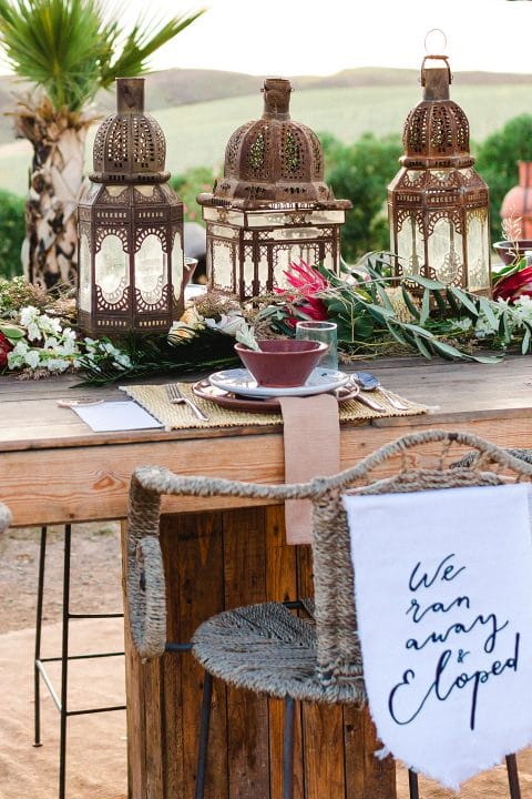 Hochzeitswahn - Ein rustikaler Hochzeitstisch im Freien mit verzierten Metalllaternen, üppigen Blumengirlanden und einem handgeschriebenen „Wir sind weggelaufen und durchgebrannt“-Schild, das ein romantisches, unkonventionelles Ambiente inmitten einer pastoralen Kulisse hervorruft.