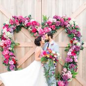 Hochzeitswahn - Ein Paar teilt einen Kuss, umrahmt von einem herzförmigen Blumenarrangement auf einer Holzkulisse, der Romantik bei ihrer Hochzeitsfeier verkörpert.