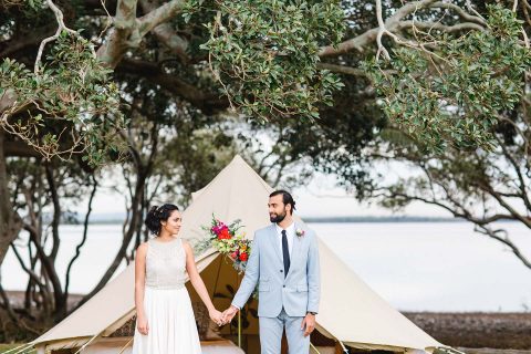 Hochzeitswahn - Ein Paar in Hochzeitskleidung hält Händchen vor einem Zelt aus Segeltuch unter einem weitläufigen Baum und strahlt eine ruhige, rustikale Eleganz inmitten der Natur aus.