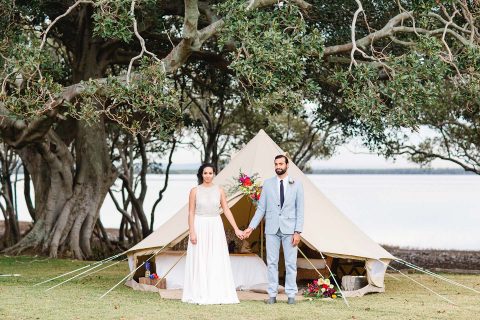 Hochzeitswahn - Ein Paar in Hochzeitskleidung hält Händchen vor einem stilvollen, mit Blumen geschmückten Glockenzelt aus Segeltuch vor einer ruhigen Kulisse am Seeufer mit üppigen Bäumen.
