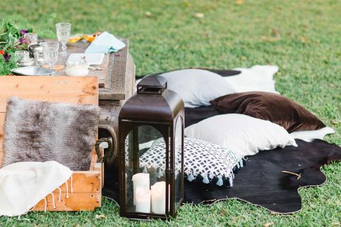 Hochzeitswahn - Ein gemütliches Picknick im Freien mit einem rustikalen Holzkistentisch, bequemen Kissen, einer Laterne mit Kerzen und einem eleganten Glas Wein auf einem üppig grünen Rasen.