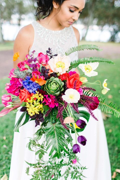 Hochzeitswahn - Eine strahlende Braut in einem eleganten weißen Kleid hält einen farbenfrohen Blumenstrauß in der Hand, der vor der grünen Kulisse eines ruhigen Parks einen atemberaubenden visuellen Effekt erzeugt.
