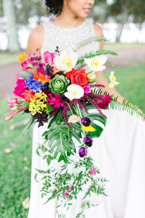 Hochzeitswahn - Eine Braut in einem weißen Kleid hält einen farbenfrohen Blumenstrauß mit einer bunten Mischung aus Blumen und Grün in der Hand, der ihr lächelndes Gesicht teilweise verdeckt, vor einem weichen, natürlichen Hintergrund.