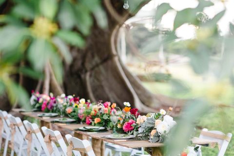 Hochzeitswahn - Ein Essbereich im Freien unter einem großen Baum mit einem rustikalen Holztisch, der mit farbenfrohen Blumenaufsätzen und eleganten Gedecken geschmückt ist, ergänzt durch weiße Klappstühle, alles inmitten üppiger Vegetation.