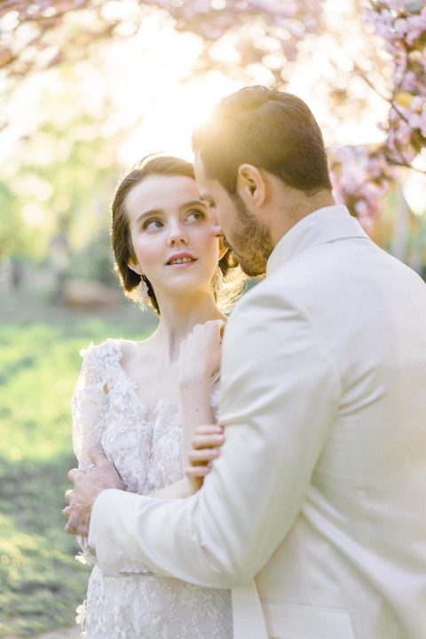 Hochzeitswahn - Eine Braut und ein Bräutigam verbringen einen intimen Moment, umgeben vom sanften Schein des Sonnenuntergangs und blühenden Bäumen, wobei ihre Umarmung Romantik und Zärtlichkeit vermittelt.