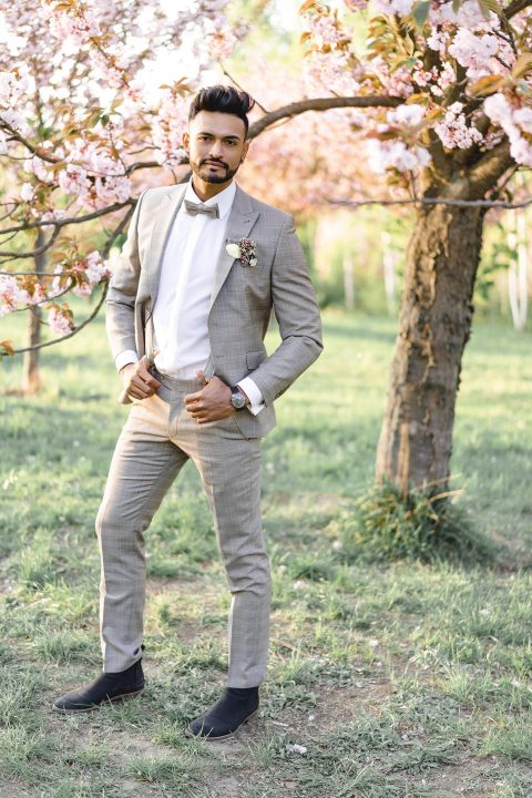 Hochzeitswahn - Ein eleganter Herr in einem stilvollen beigen Anzug und einer Fliege steht selbstbewusst unter Kirschblüten und strahlt Eleganz und Frühlingsgefühle aus.