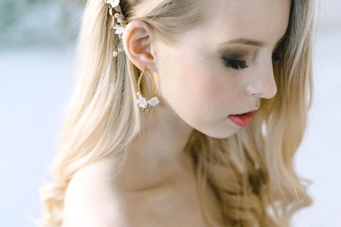 Hochzeitswahn - Ein Porträt einer Frau mit wehmütigem Gesichtsausdruck, die vor einem weichen, hellen Hintergrund zarte Blumenaccessoires in ihrem Haar und elegante Ohrringe zur Schau stellt.