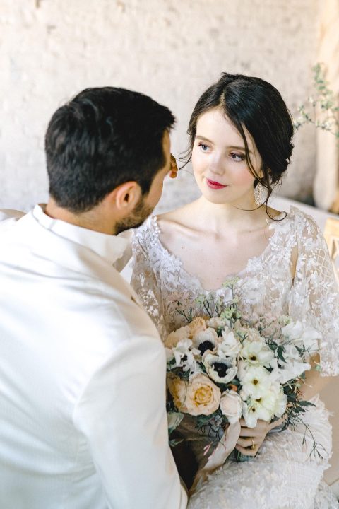 Hochzeitswahn - Eine Braut in einem Spitzenhochzeitskleid hält einen Blumenstrauß in der Hand und blickt ihren Partner in einem eleganten Anzug während eines ruhigen gemeinsamen Moments liebevoll an.