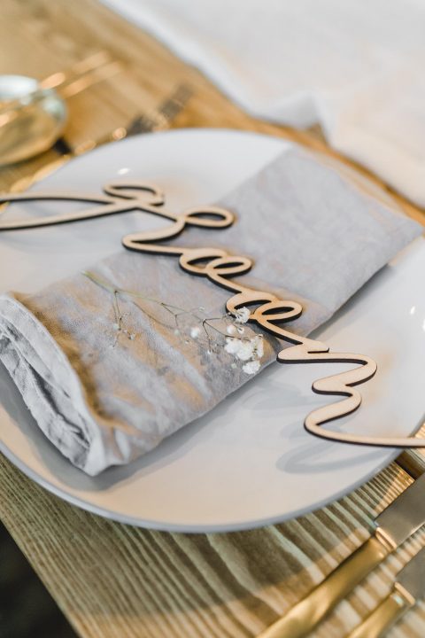 Hochzeitswahn - Eine elegante Tischdekoration mit einem weißen Teller und einer grauen Leinenserviette, garniert mit einer zarten „Prost“-Schriftzugdekoration, die eine elegante und festliche Atmosphäre beim Essen vermittelt.