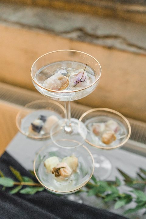 Hochzeitswahn - Eine Reihe eleganter Martini-Gläser präsentieren kunstvoll verschiedene Gourmet-Eiskugeln, jeweils geschmückt mit zarten essbaren Blumen, präsentiert auf einem Metalltablett vor einem rustikalen Holzhintergrund.