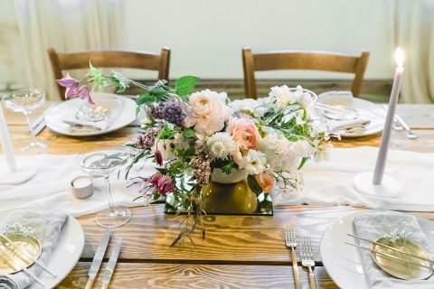Hochzeitswahn - Ein elegant gedeckter Esstisch mit einem üppigen Blumenaufsatz in einer Messingvase, flankiert von zwei hohen brennenden Kerzen, mit feinem Porzellan und Silberbesteck auf einer frischen weißen Leinentischdecke.