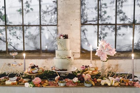 Hochzeitswahn - Ein eleganter Hochzeits-Desserttisch, geschmückt mit einer dreistöckigen Torte, Blumendekorationen, einer Auswahl an Früchten, Gebäck und brennenden Kerzen, die nachts am Fenster eine romantische Atmosphäre schaffen.