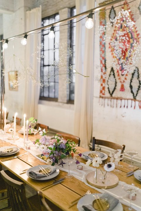 Hochzeitswahn - Ein elegant gedeckter Esstisch mit einer Mischung aus rustikalem Charme und moderner Ästhetik, mit warmem Kerzenlicht, frischen Blumenarrangements und schicken hängenden Glühbirnen; Schaffen Sie eine gemütliche und zugleich elegante Atmosphäre für ein Treffen.