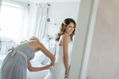 Hochzeitswahn - Eine strahlende Braut in einem Spitzenbrautkleid erhält in einem ruhigen, sonnendurchfluteten Raum kurz vor der Zeremonie den letzten Schliff von ihrer Brautjungfer.