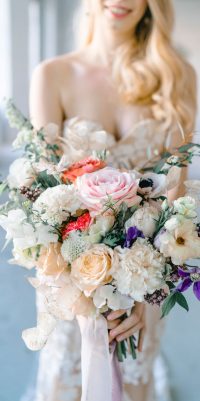 Hochzeitswahn - Eine Braut in einem trägerlosen Kleid lächelt sanft und hält einen großen, eleganten Blumenstrauß aus rosa Rosen, weißen Blüten und Grünpflanzen, akzentuiert mit Bändern, die eine romantische Atmosphäre schaffen.