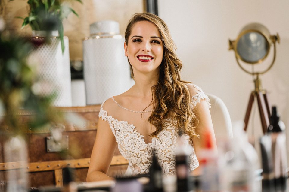Hochzeitswahn - Eine lächelnde Frau in einem Spitzenbrautkleid sitzt an einem Schminktisch, umgeben von Make-up-Produkten, und ein Spiegel spiegelt ihren freudigen Gesichtsausdruck wider. Magical-Homes