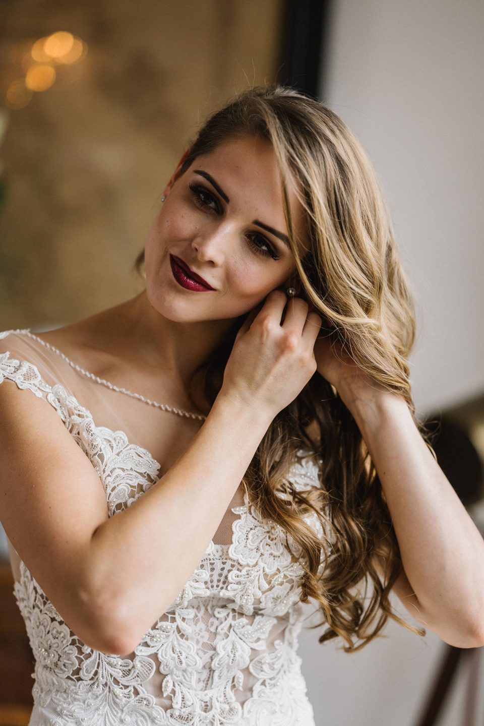 Hochzeitswahn - Eine Frau mit langem, welligem Haar und einem detailreichen Spitzenbrautkleid posiert im Innenbereich, lächelt sanft und berührt sanft ihren Ohrring. Der Hintergrund ist leicht unscharf. Magical-Homes