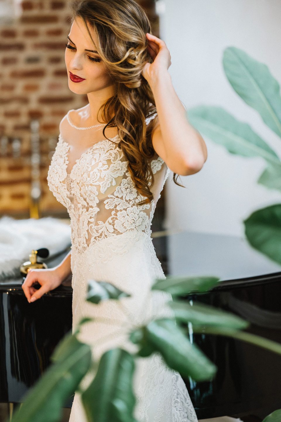 Hochzeitswahn - Eine Frau in einem eleganten Spitzenbrautkleid berührt ihr lockiges Haar, während sie neben einem schwarzen Klavier steht. Im Vordergrund sind grüne Pflanzen zu sehen, im Hintergrund eine Backsteinmauer. Magical-Homes
