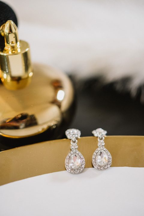 Hochzeitswahn - Ein Paar elegante Tropfenohrringe mit Diamantakzenten auf einer goldenen reflektierenden Oberfläche mit weichgezeichnetem Hintergrund mit dunklen und weißen Texturen. Magical-Homes