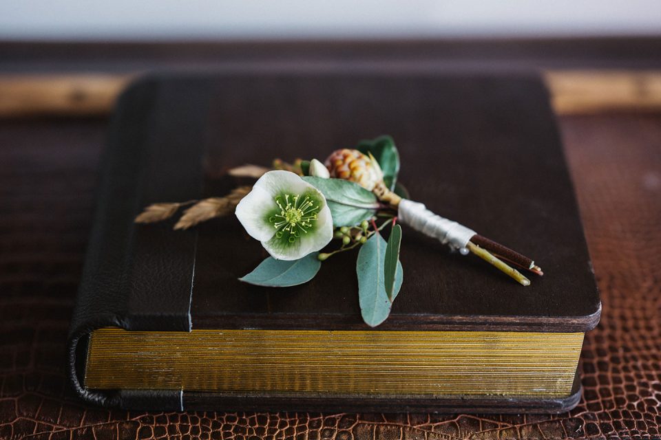 Hochzeitswahn - Ein geschlossenes, abgenutztes gebundenes Buch mit Goldschnitt auf Holzoberfläche, geschmückt mit einem kleinen Strauß aus einer weißen Blume, getrockneten Blättern und Zweigen, zusammengebunden mit einer weißen Schleife. Magical-Homes