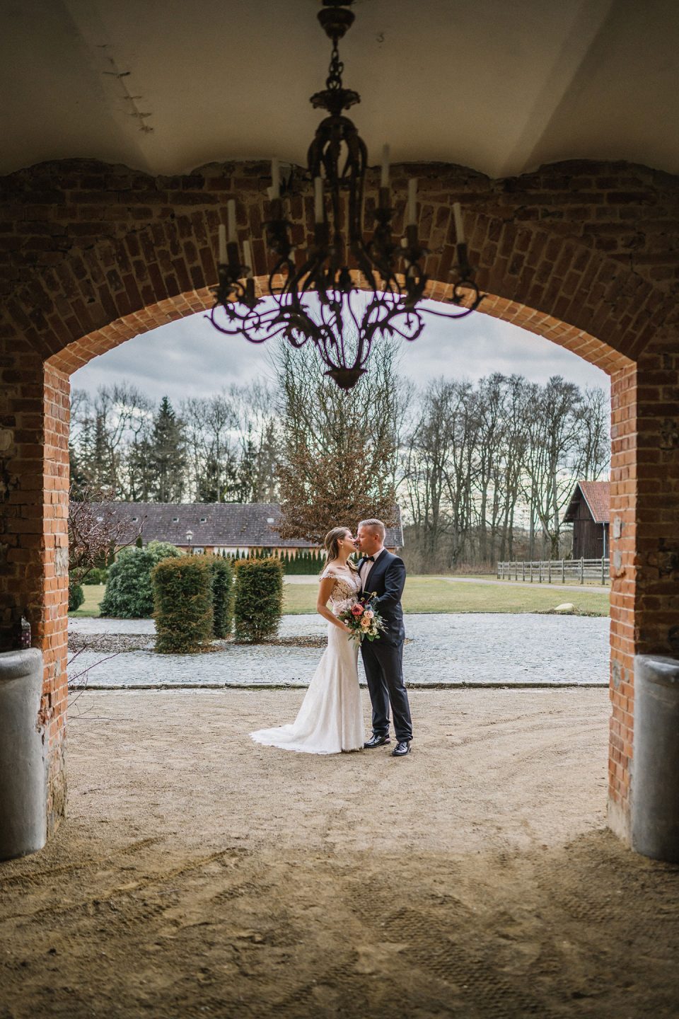 Hochzeitswahn - Eine Braut und ein Bräutigam umarmen sich unter einem gewölbten Backsteineingang, mit Blick auf einen üppigen Park im Hintergrund. Über ihnen hängt ein kunstvoller Kronleuchter. Magical-Homes