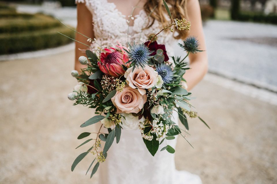 Hochzeitswahn - Eine Braut in einem Spitzenkleid hält einen farbenfrohen Strauß mit Rosen und exotischen Blumen vor einer ruhigen Wasserkulisse. Nur der Strauß und ein Teil ihres Kleides sind sichtbar. Magical-Homes