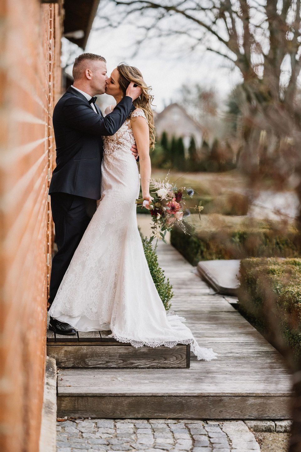 Hochzeitswahn - Braut und Bräutigam verbringen einen intimen Moment neben einem Backsteingebäude. Sie stehen auf einem Holzweg mit viel Grün am Rand. Die Braut trägt ein Spitzenkleid und wird vom Bräutigam im Anzug umarmt. Magical-Homes