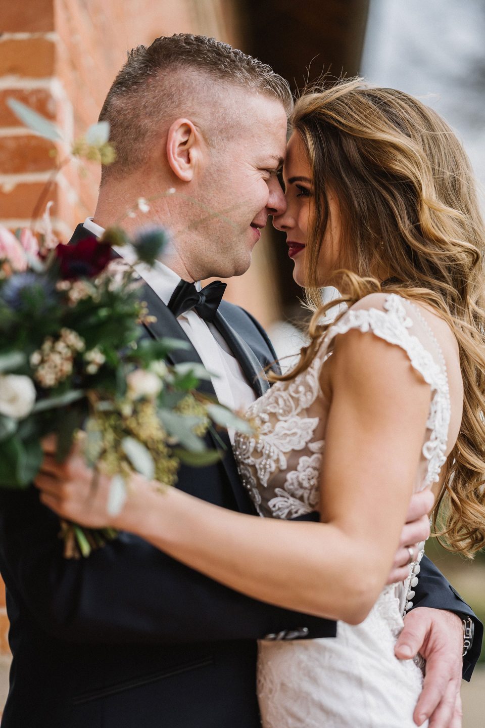 Hochzeitswahn - Braut und Bräutigam teilen einen intimen Moment im Freien. Ihre Stirnen berühren sich und ihre Augen sind geschlossen. Die Braut hält einen Blumenstrauß und beide tragen formelle Hochzeitskleidung. Magical-Homes