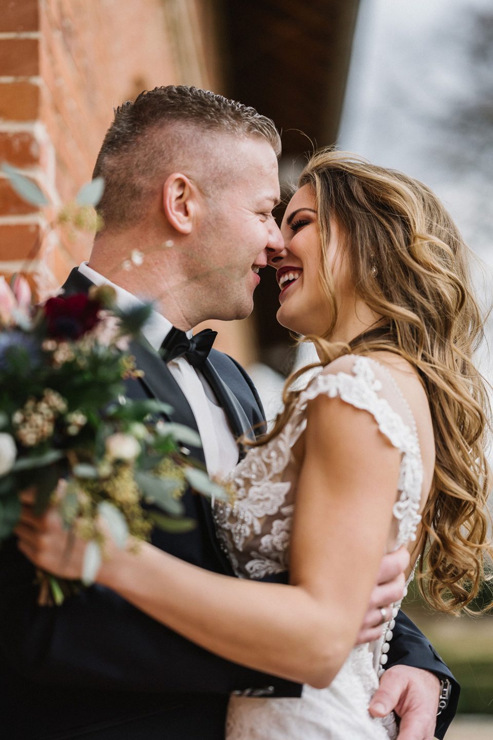 Hochzeitswahn - Eine freudige Braut und ein Bräutigam umarmen sich, ihre Nasen berühren sich, während sie lächeln und einen Strauß dunkler Blumen in den Händen halten. Der Bräutigam trägt einen schwarzen Smoking und die Braut ein detailreiches Spitzenkleid. Magical-Homes