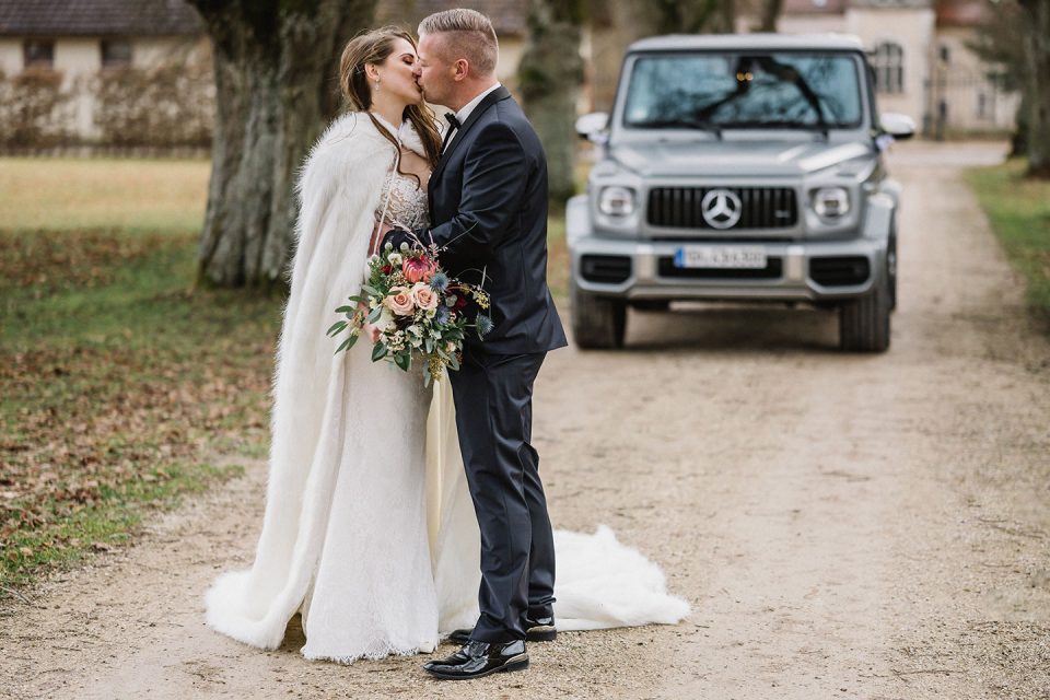 Hochzeitswahn - Braut und Bräutigam küssen sich auf einer unbefestigten Straße. Die Braut trägt einen langen weißen Mantel und hält einen Blumenstrauß in der Hand. Im Hintergrund parkt ein Luxus-SUV. Magical-Homes