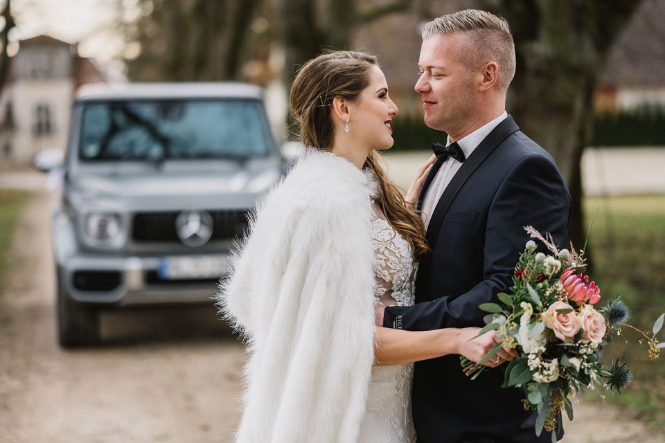 Hochzeitswahn - Eine elegant gekleidete Braut und ein Bräutigam verbringen einen freudigen Moment im Freien, halten Händchen und schauen sich neben einem geparkten Luxusauto an. Die Braut hält einen Blumenstrauß und die Szenerie vermittelt den Eindruck einer heiteren, romantischen Atmosphäre. Magical-Homes