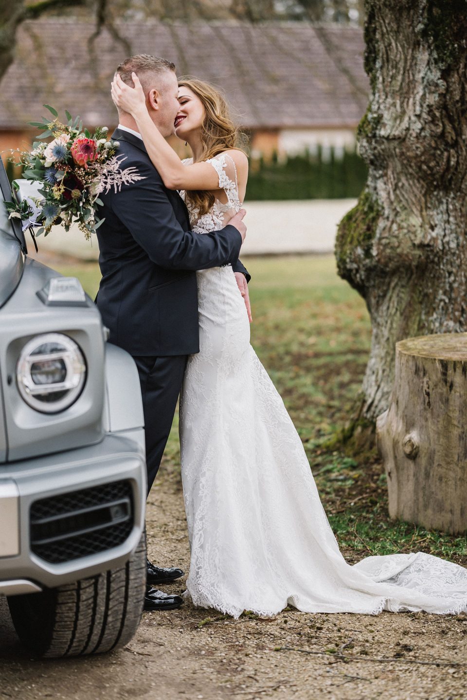 Hochzeitswahn - Eine Braut und ein Bräutigam küssen sich liebevoll neben einem geparkten Auto unter einem großen Baum, wobei die Braut einen Blumenstrauß hält. Die Braut trägt ein Spitzenbrautkleid und der Bräutigam einen dunklen Anzug. Magical-Homes