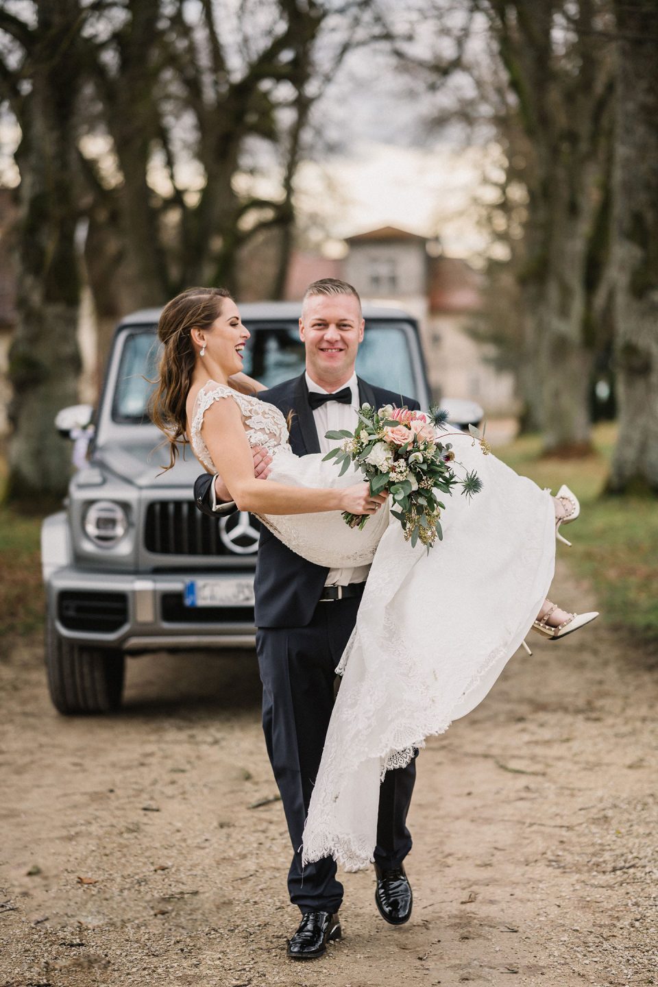 Hochzeitswahn - Ein Bräutigam in schwarzem Anzug und Fliege hebt die Braut in einem weißen Spitzenkleid vom Boden hoch. Beide lächeln und posieren vor einem schwarzen Geländewagen mit Bäumen und einem bewölkten Himmel im Hintergrund. Magical-Homes
