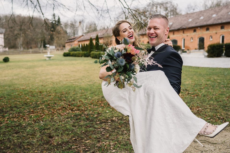 Hochzeitswahn - Eine fröhliche Braut, die in den Armen des Bräutigams getragen wird, lacht herzlich und hält einen Blumenstrauß in der Hand. Sie befinden sich im Freien vor einer Kulisse aus blattlosen Bäumen und historischen Gebäuden. Magical-Homes