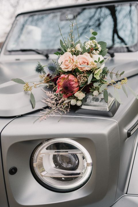 Hochzeitswahn - Ein Blumenarrangement mit rosa Rosen und einer roten Protea auf der Motorhaube eines silbernen Autos, platziert neben dem rechten Scheinwerfer, vor einem unscharfen Hintergrund aus Bäumen. Magical-Homes