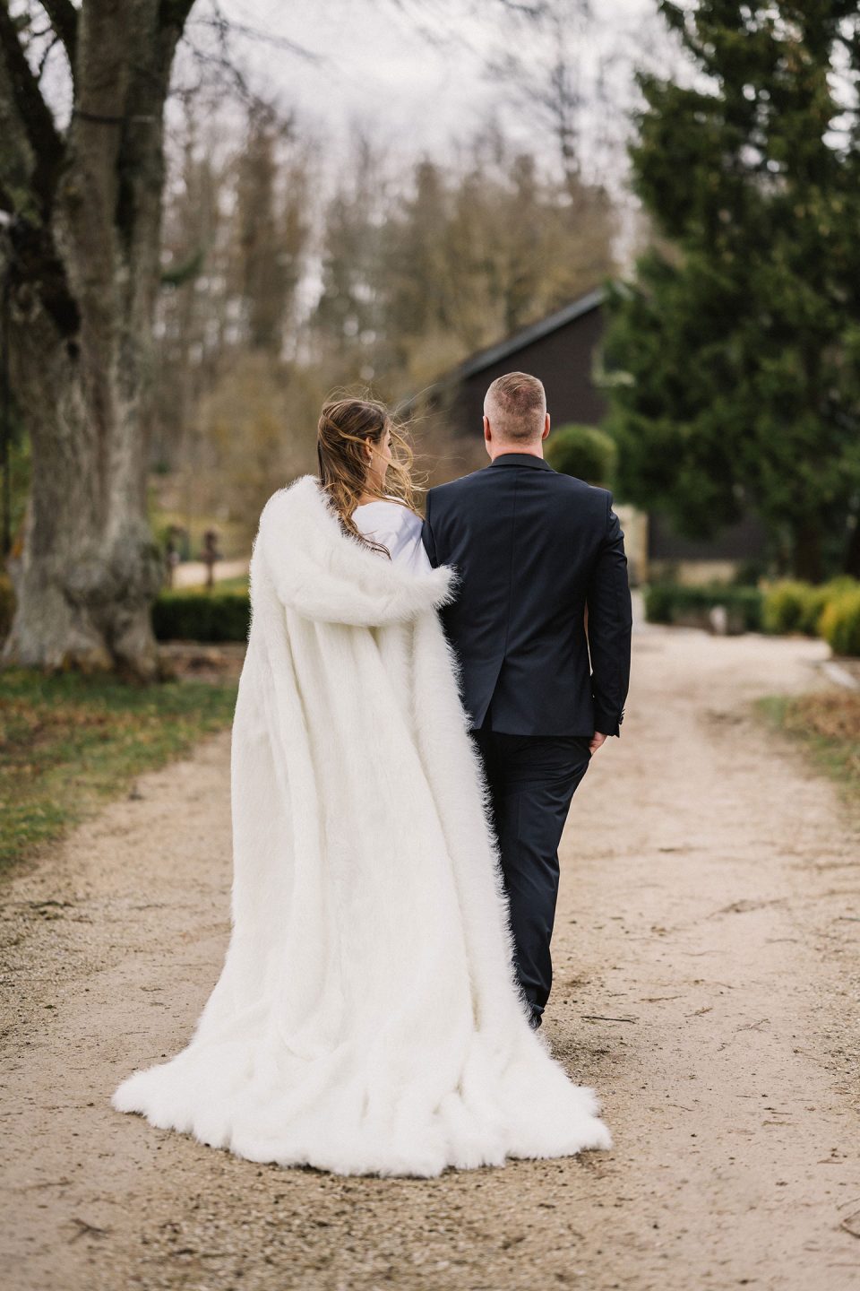 Hochzeitswahn - Braut und Bräutigam gehen Hand in Hand einen Kiesweg entlang. Die Braut trägt ein elegantes weißes Kleid mit einem langen Pelzmantel und der Bräutigam einen klassischen schwarzen Anzug. Magical-Homes