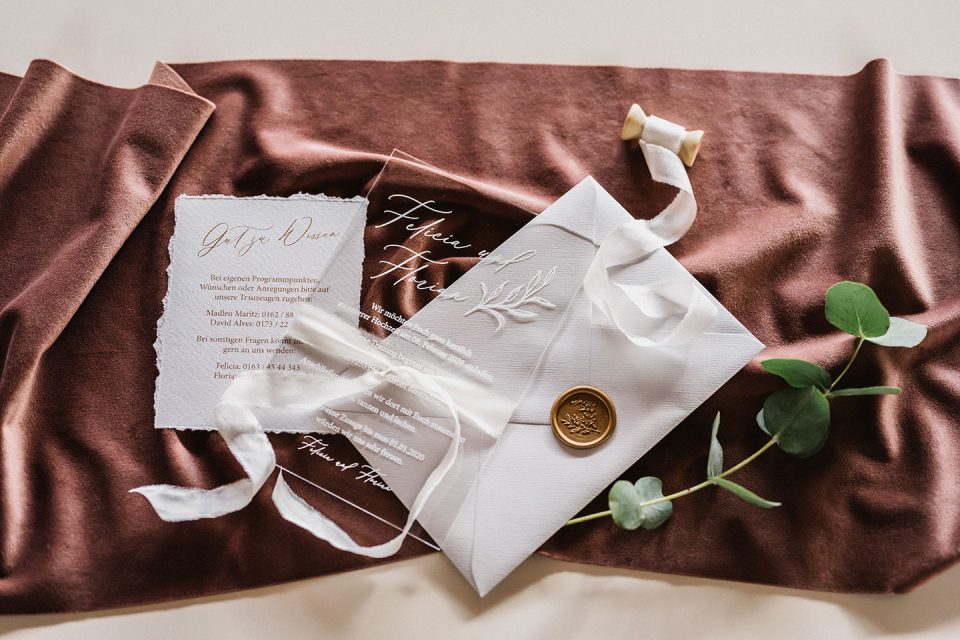 Hochzeitswahn - Elegante Hochzeitseinladung und Antwortkarte mit weißer Kalligrafie auf durchscheinendem Papier, zusammengebunden mit einem Seidenband, einem Wachssiegel und grünen Blättern auf einem satten burgunderfarbenen Stoffhintergrund. Magical-Homes