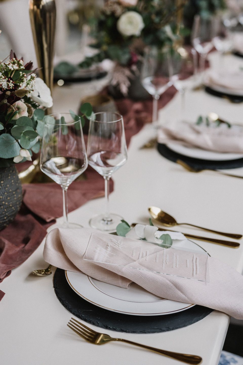 Hochzeitswahn - Elegant gedeckter Esstisch mit schwarzen Tellern, goldenem Besteck und rosa Servietten, verziert mit einer dekorativen weißen Menükarte. Weingläser aus Kristall und ein Blumengesteck runden den anspruchsvollen Look ab. Magical-Homes